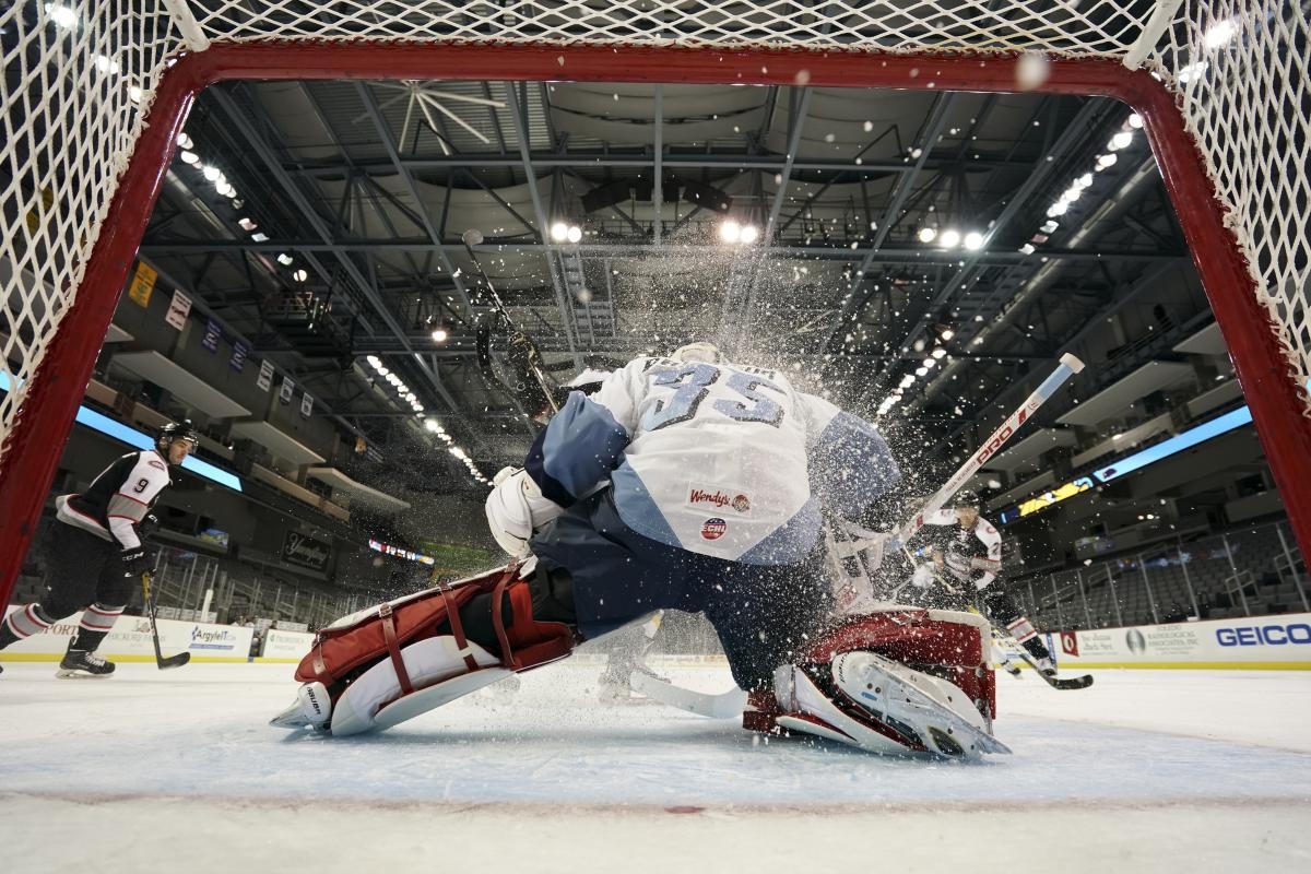 View from the back of an ice hockey net as the goalkeeper dives to stop a shot from opponents