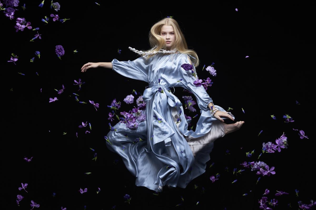 Female model in flowing dress, jumping and surrounded by purple flowers, illuminated against a black background 