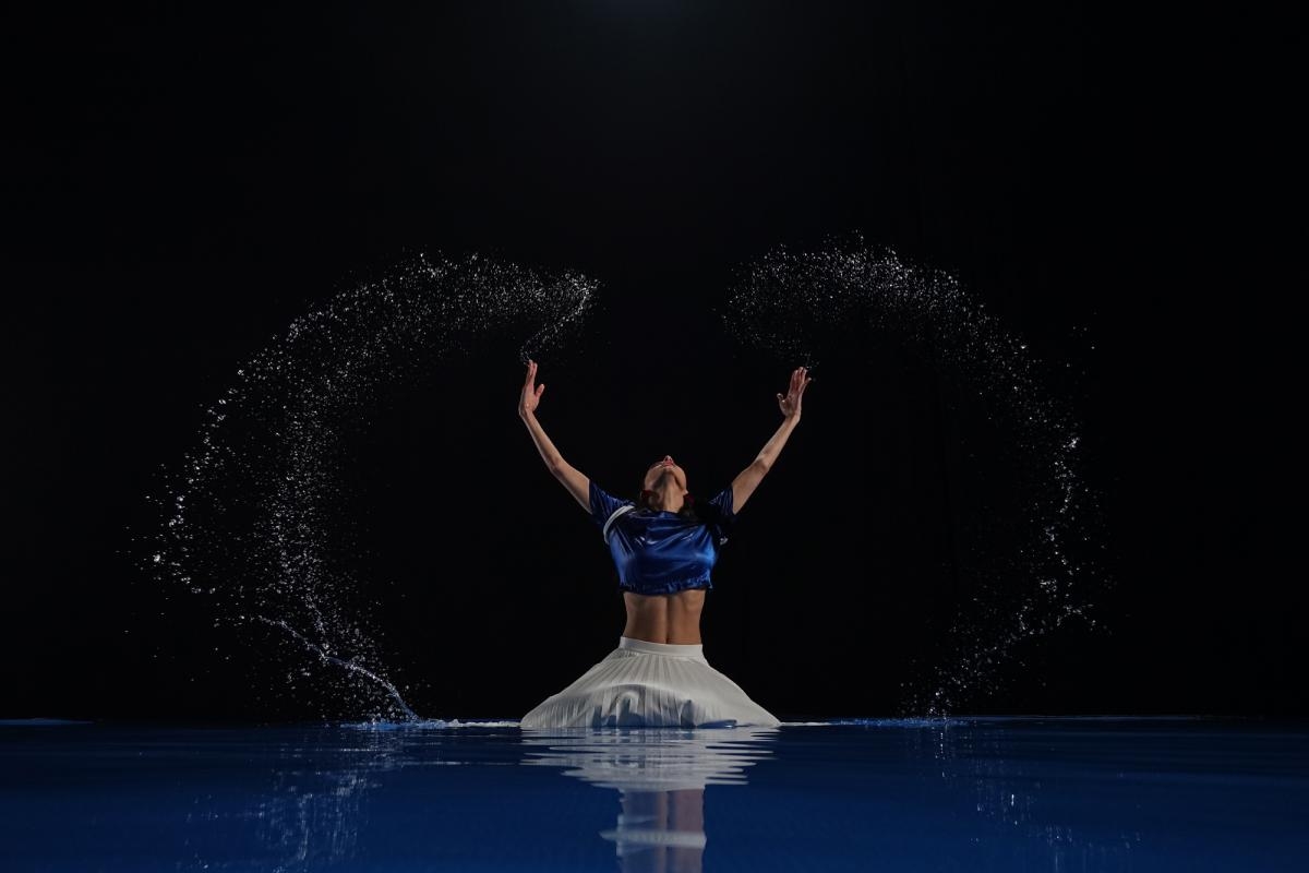 Female dancer kneeling in water, arms stretched above her head, illuminated against a black background