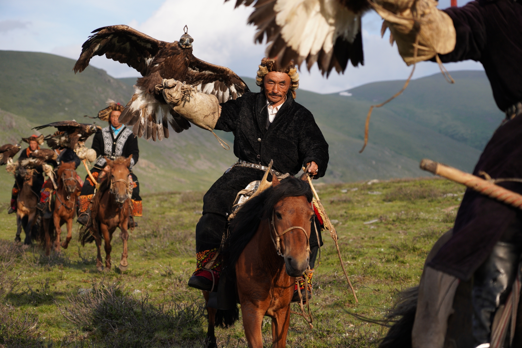 Horse riders crossing grassy, mountainous terrain with falcons on their aims