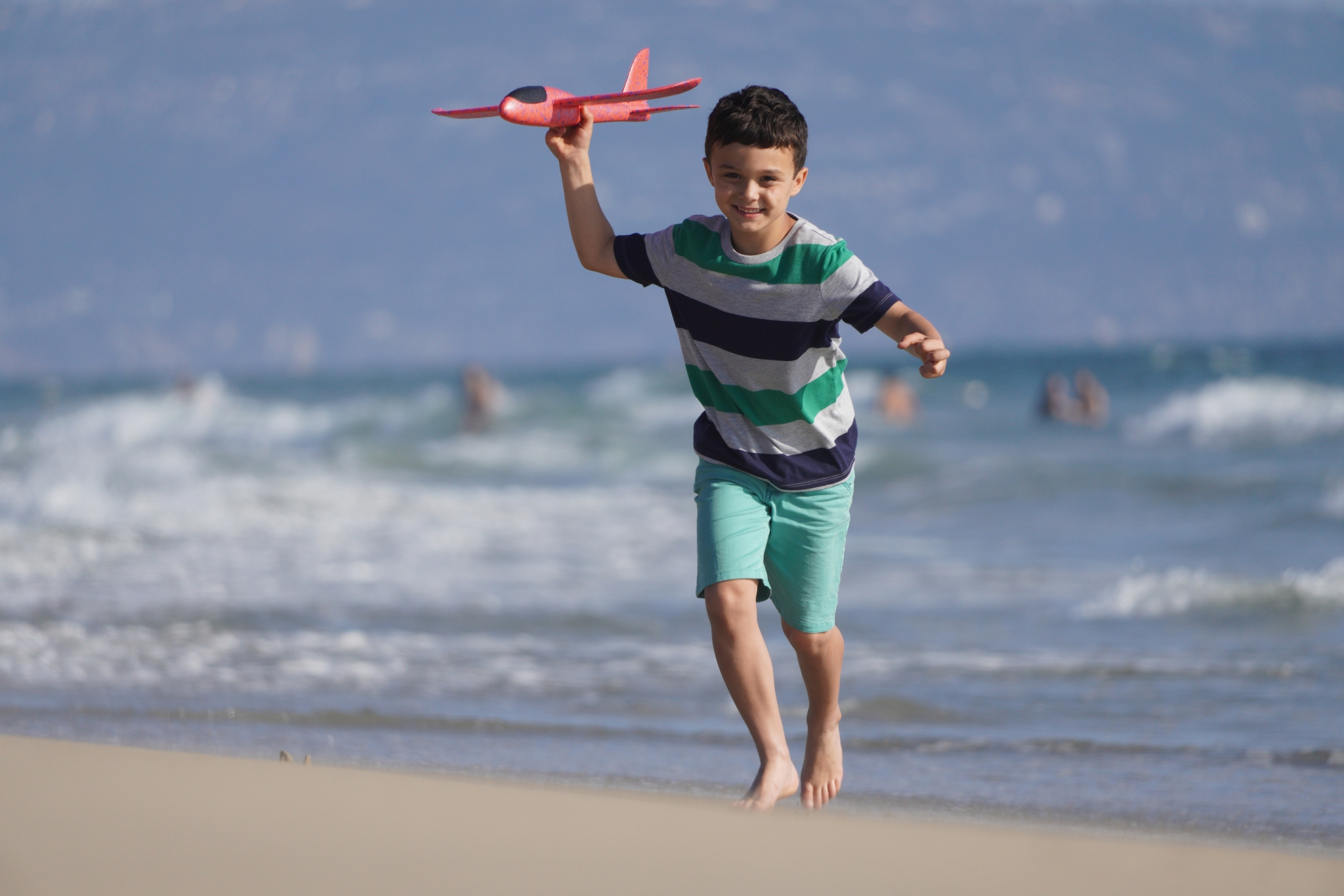Young boy running on beach and playing with red toy aeroplane with the sea in a bokeh background