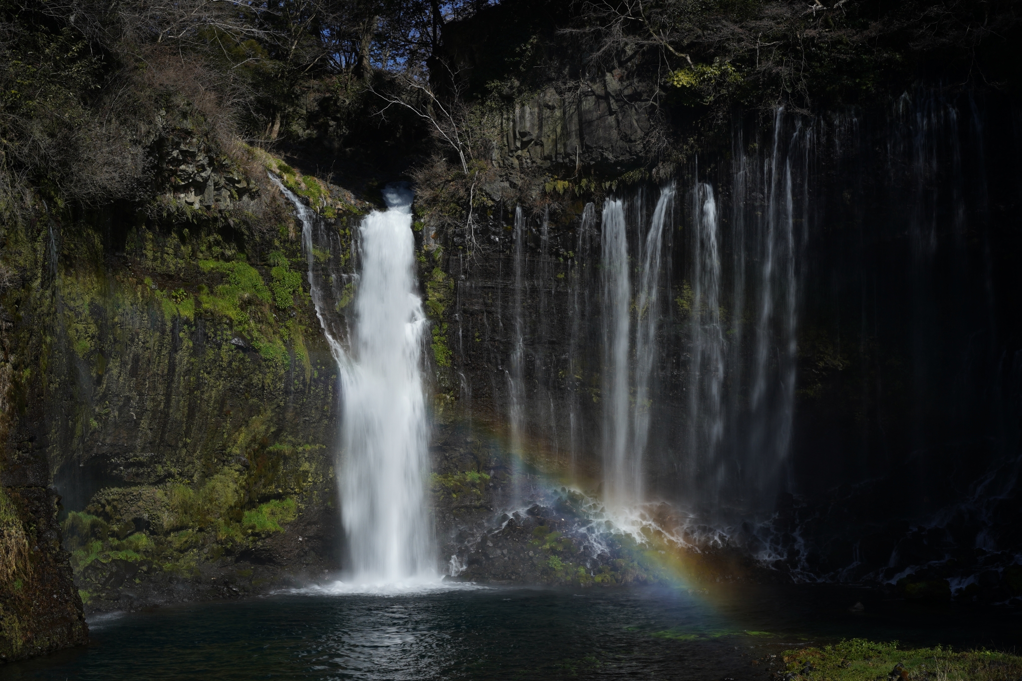 Wide waterfall with forested surroundings and a rainbow visible in the water spray