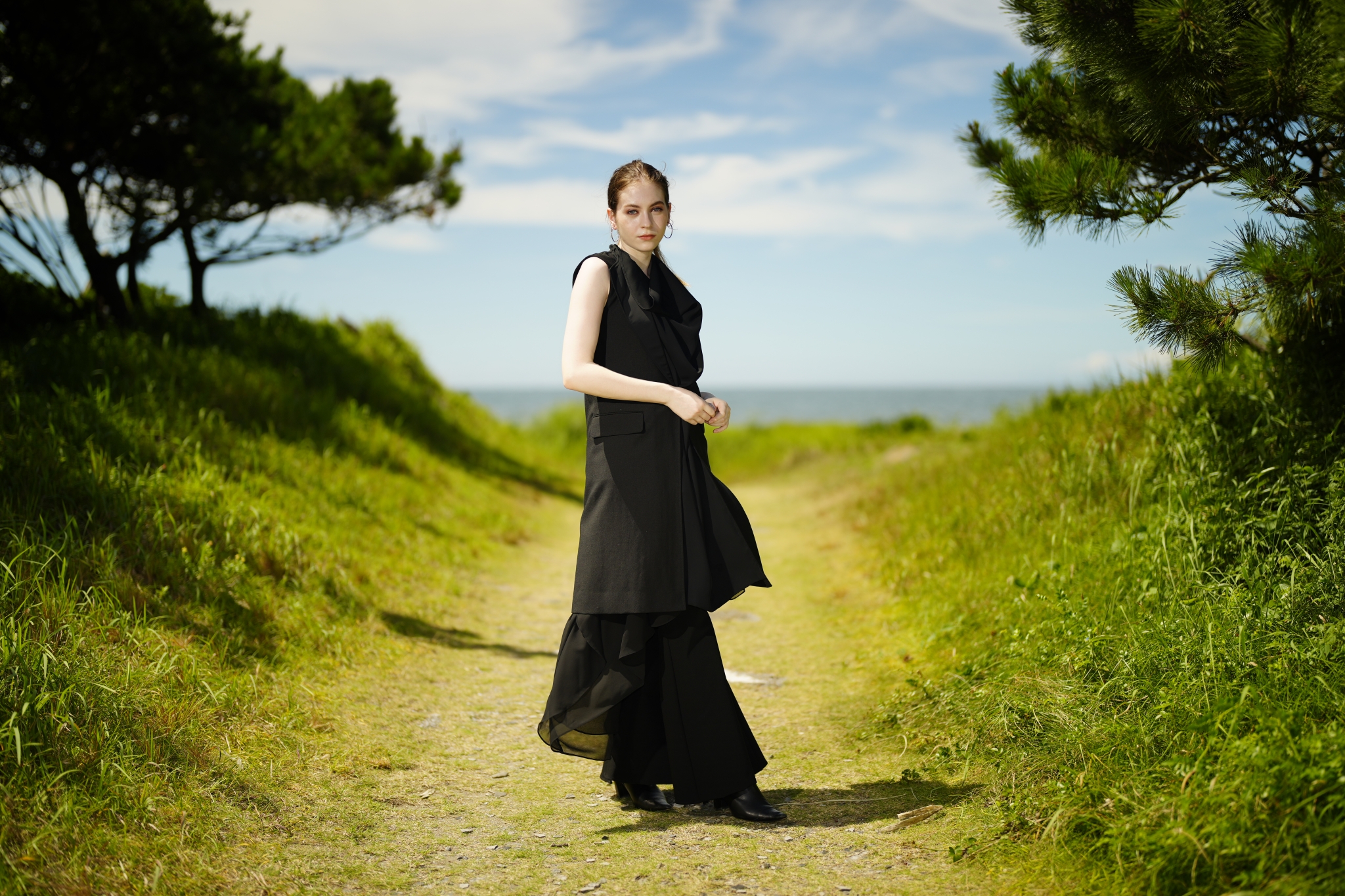 Female model in a long black dress stood on a grassy path leading to the ocean