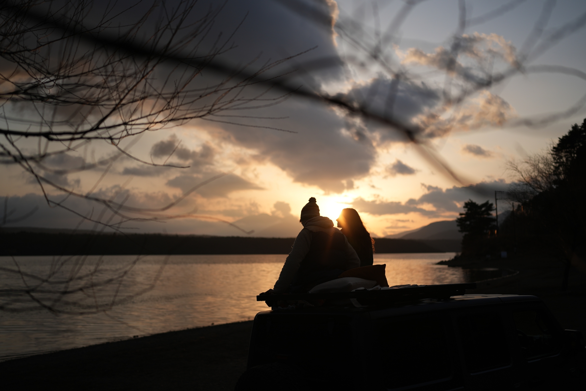 A couple sat by a lake, silhouetted against the setting sun