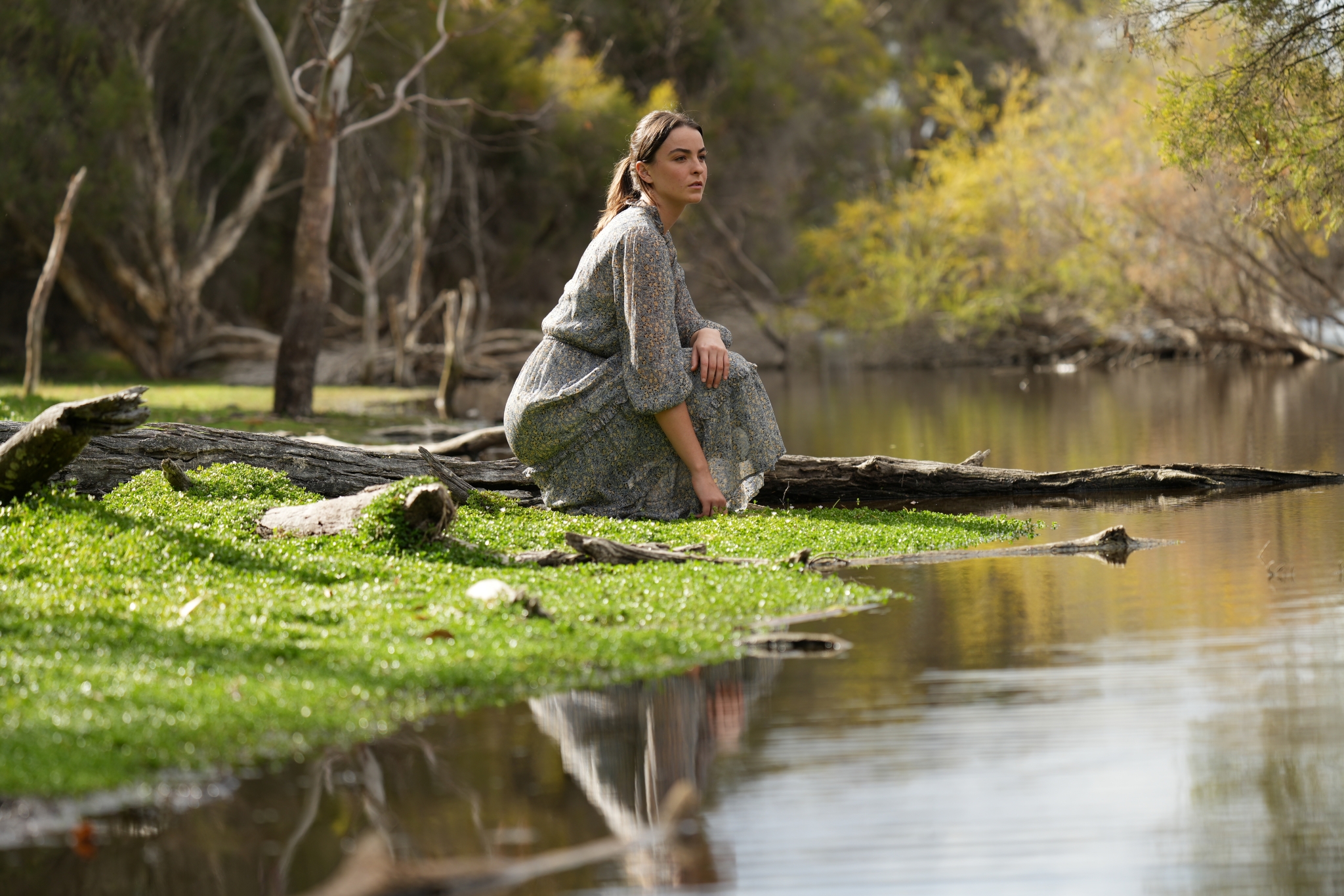 Female model sat on a grassy riverbank in a forest