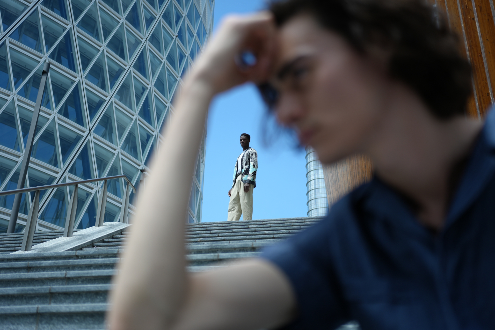 A male model stood at the top of stairs, framed by the arm of a male model in bokeh foreground