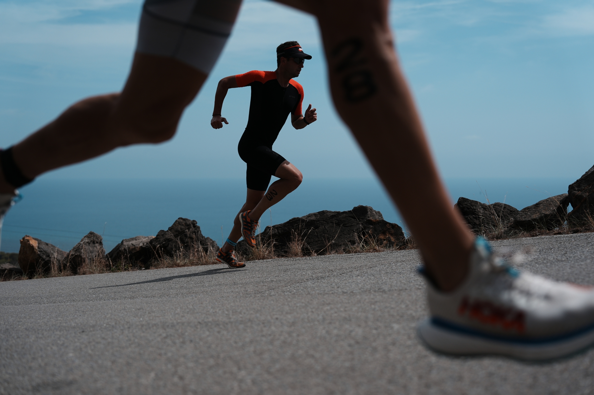 Male runner running along a road by the sea, viewed from between another runner's legs in bokeh foreground