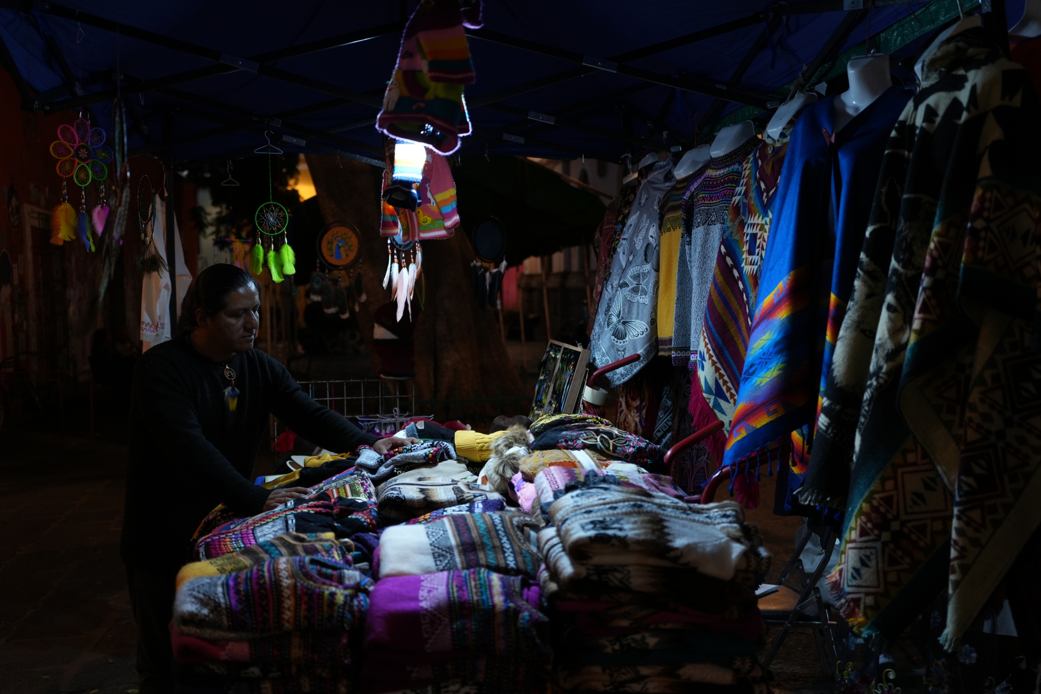 A man supervises a scarf, ties and garment stall lit by a single bulb