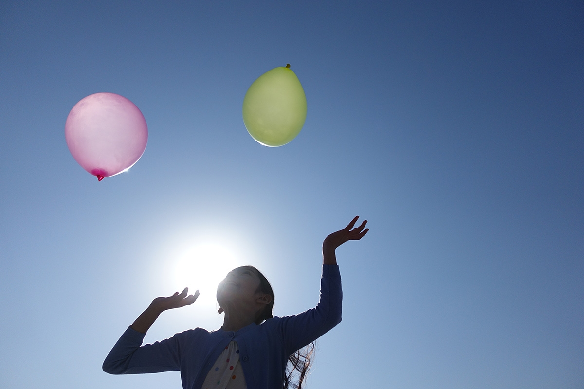 Girl silhouetted against sun releasing red and yellow balloons