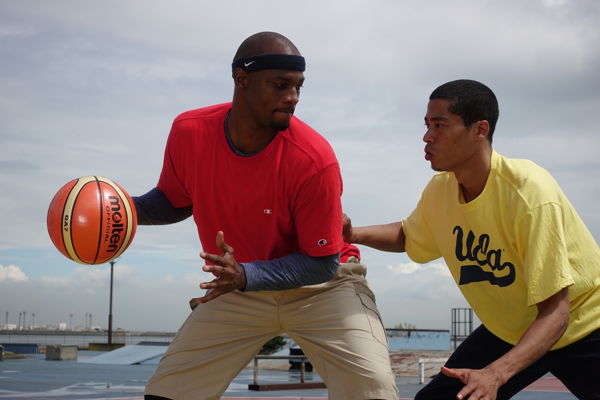 Two men playing basketball in outdoor location, one defending against the other