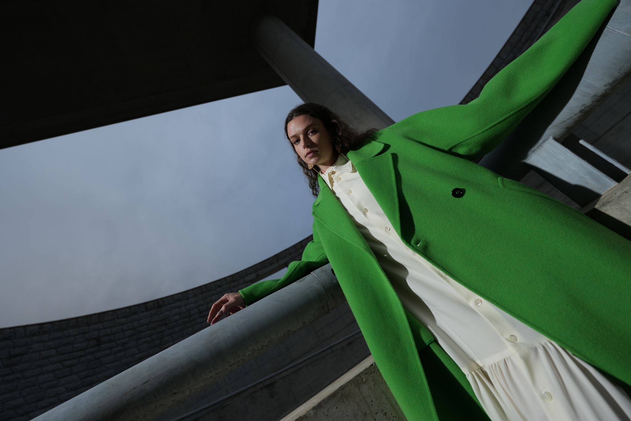 A low-angle photo of a woman in a green coat leaning on her back and looking down. Blue skies and modern architecture are above her head.