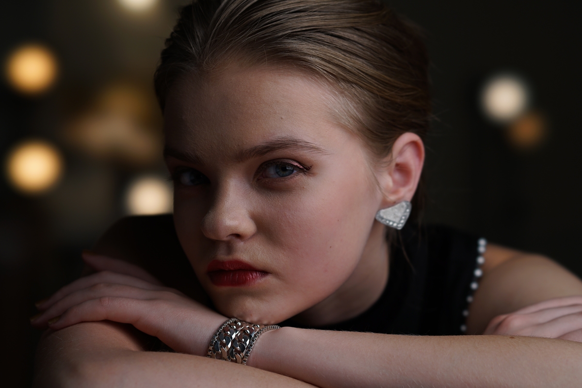 Close-up portrait of woman looking into camera resting head on arms with lights in dark background bokeh