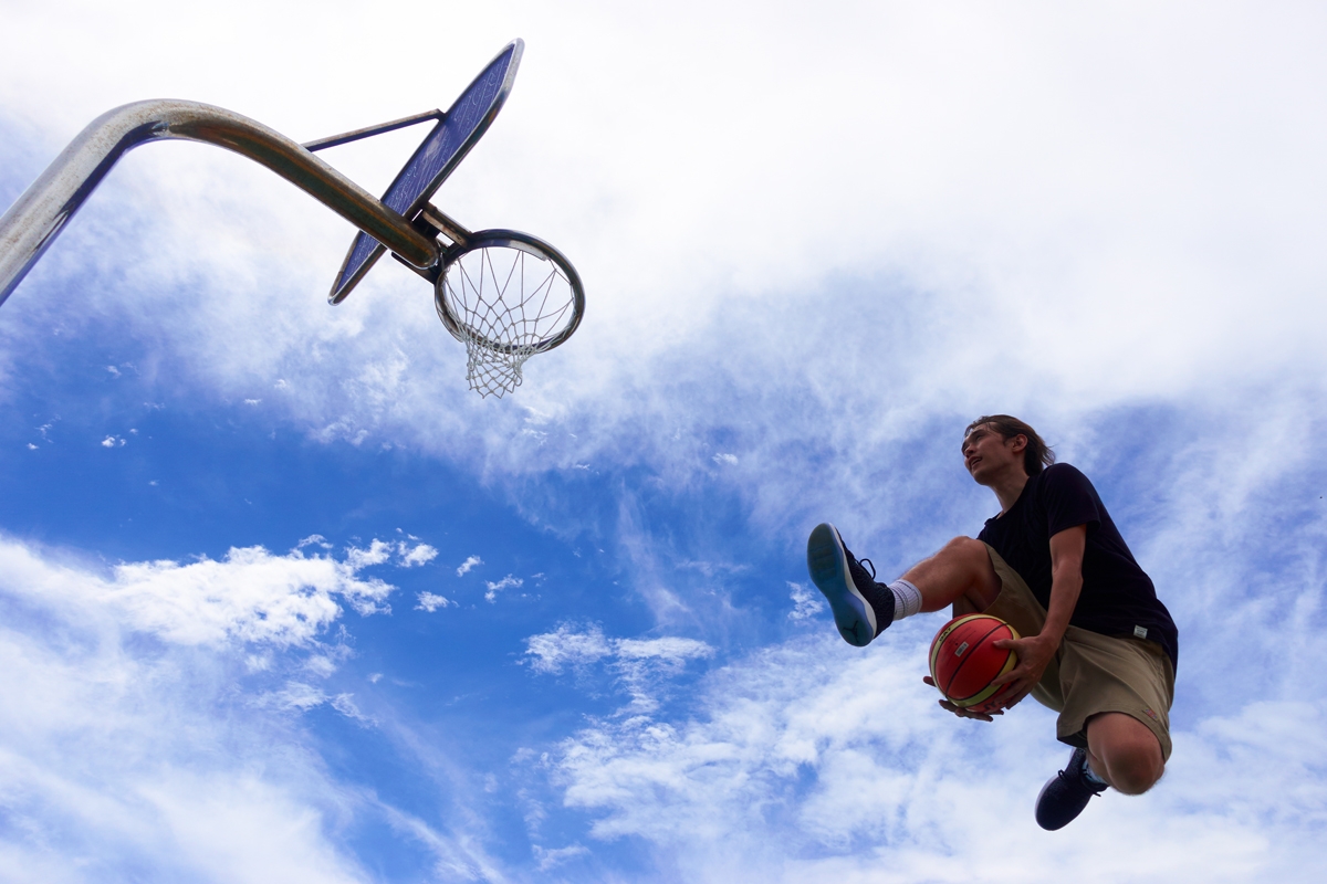 Basketball player jumping toward hoop in outdoor location, passing the ball under his leg